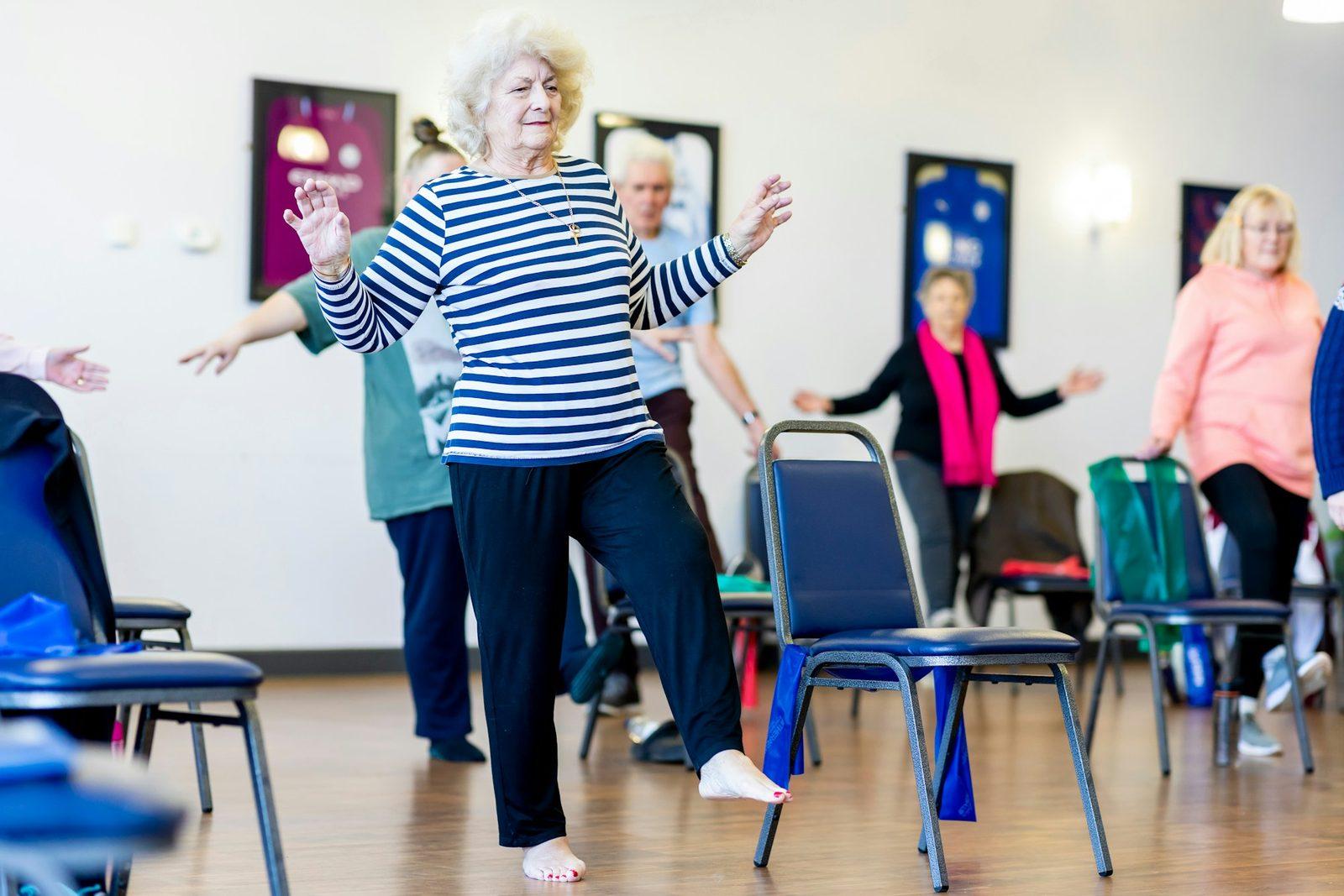 An older woman practising a single-leg balance exercise with chair support.